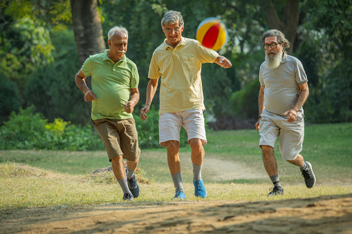 Active senior male friends playing with soccer ball on grassy field against trees in garden during weekend