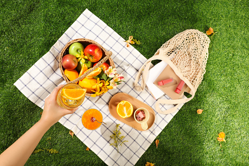 Picnic basket with fruits, flowers, lipsticks and book on the white checkered mat in the green park background. Hand of woman is holding a cool juice cups