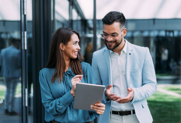 Business people using digital tablet outdoors. Shot of two confident colleagues walking and talking next to an office building. Businessman and businesswoman in meeting using digital tablet and discussing business strategy. Creative business persons discussing new project and sharing ideas while walking in front of their office building. business people stock pictures, royalty-free photos & images