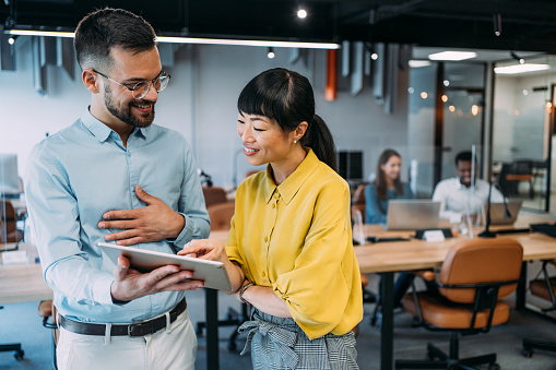 Shot of two coworkers having a discussion in modern office. Businessman and businesswoman in meeting using digital tablet and discussing business strategy. Confident business people working together in the office. Creative business persons discussing new project and sharing ideas in the workplace while their colleagues working in background.