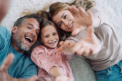 Above view of beautiful young parents and their cute little daughter looking at camera and smiling while lying on carpet in the living room at home.