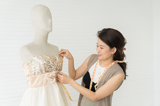 Asian female dressmaker with hanging measuring tape getting dressed dummy mannequin in tailor shop. Tailor shop. Garment, design, fashion, dressmaking concept