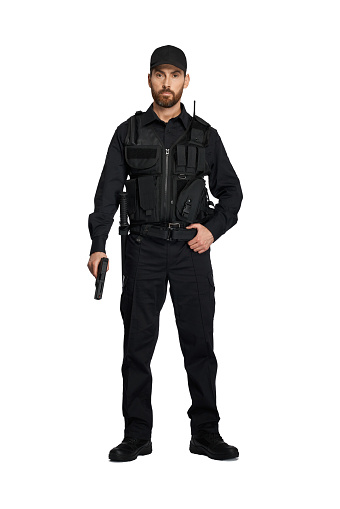 Confident police officer in black uniform posing with weapon indoors. Front view of bearded policeman with hand on belt, holding police handgun, isolated on white. Concept of work, profession, weapon.