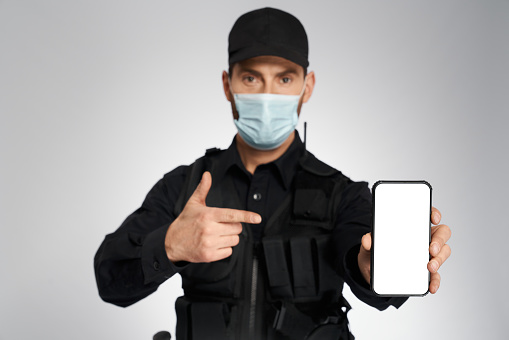 Male policeman in protective mask pointing to empty cell phone's screen. Portrait of caucasian cop gesturing, while looking camera, on gray studio background. Concept of safety, calling, quarantine.