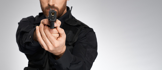 Looking down barrel of anonymous police officer's sidearm in studio. Crop view of unrecognizable constable aiming, by pointing handgun to camera, isolated on gray. Concept of danger, work, weapon.