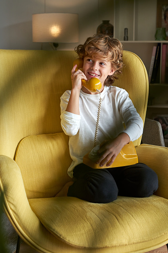 Full body of positive boy with curly hair talking on retro landline telephone in armchair in light room with blurred plant