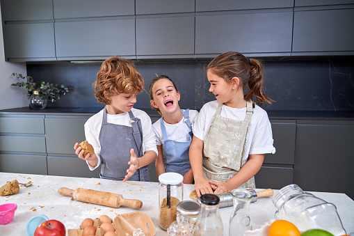 Cheerful children in aprons kneading dough for cookies at messy table with various ingredients in light modern kitchen at home
