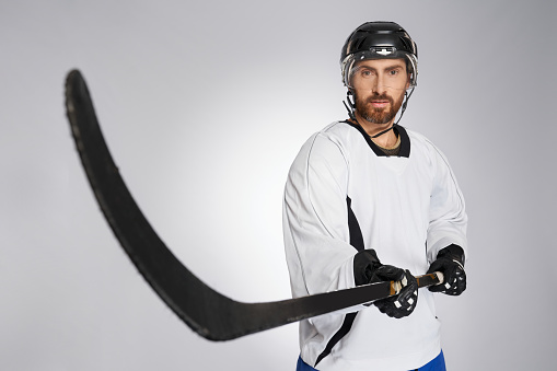 Bearded hockey player in protective shield pointing black stick into camera. Close up view of caucasian male ice hockey sportsman in uniform performing slap shot, isolated on gray. Concept of sport.