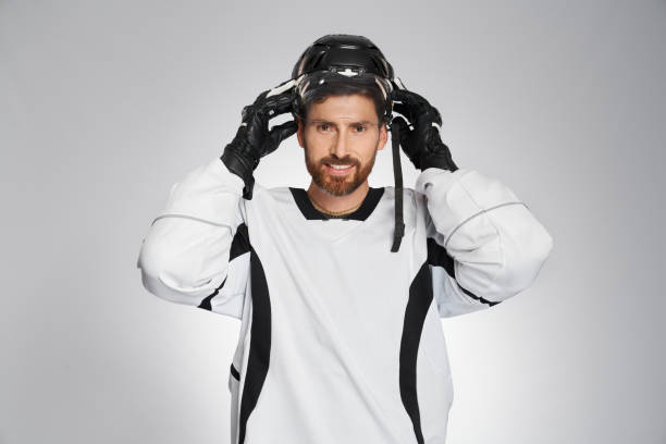 Smiling bearded ice hockey player putting on protective helmet indoors. Portrait of male sportsman using hockey face mask before game, isolated on gray studio background. Concept of hockey, safety.