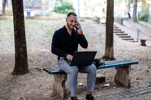 Opting for an outdoor workspace, a young businessman sits on a park bench, balancing his laptop and mobile phone effectively