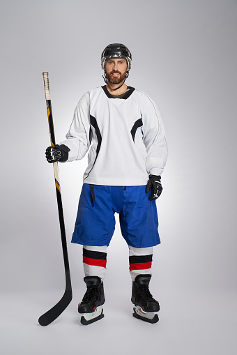 Smiling, bearded ice hockey player with stick in light studio. Front view of male hockey athlete in full gear, ready to play, isolated on gray background. Concept of winter team game.