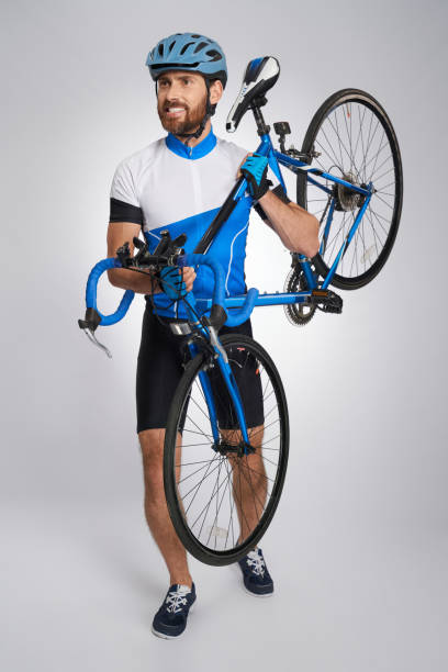 Strong, professional cyclist in helmet carrying bicycle in studio. Front view of handsome bearded athlete in cycle outfit lifting bike, isolated on grey background. Concept of sport activity, cycling.