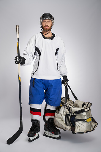 Strong bearded ice hockey player carrying stick and sport bag after game. Front view of male hockey athlete in full gear looking at camera, isolated on gray background. Concept of winter team game.
