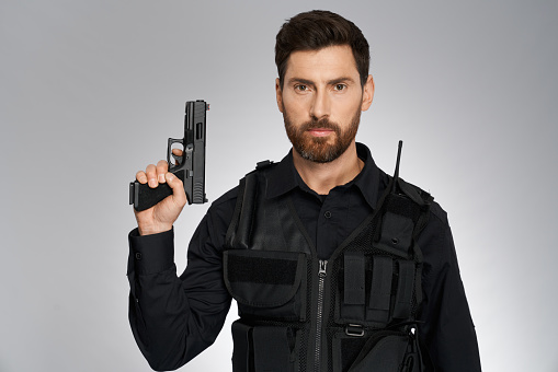 Handsome bearded cop with handgun, posing to camera. Portrait of confident policeman in body armor armed with short-barrelled gun, on gray studio background. Concept of police profession, equipment.