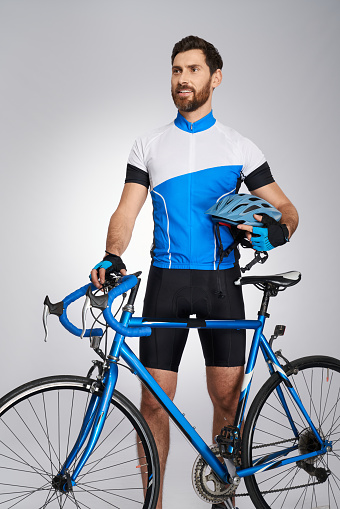 Professional cyclist with helmet and bicycle standing in studio. Front view of handsome bearded biker in cycle kit looking away, isolated on grey background. Concept of sport, cycling activity.