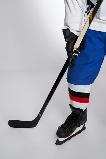 Unrecognizable male ice hockey player with stick in light studio. Crop view of strong man in full hockey equipment and black skates, isolated on gray background. Concept of winter sports, team game.