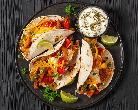 baked tex-mex chicken fajitas with mixed sweet pepper, onion, sour cream, shredded cheese and white corn tortillas on black plate on black wooden table, horizontal view, flat lay, close-up