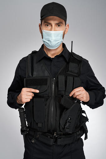Young caucasian policeman wearing protective mask looking at camera. Portrait of confident police officer in bulletproof vest posing, isolated on gray studio background. Concept of work, quarantine.