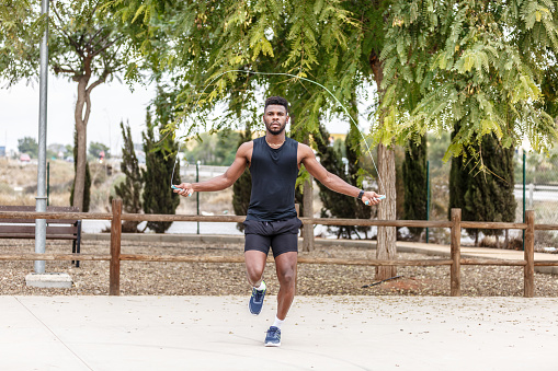 Full body of bearded African American sportsman in black activewear jumping rope against green coniferous trees in summer park during fitness training in daylight