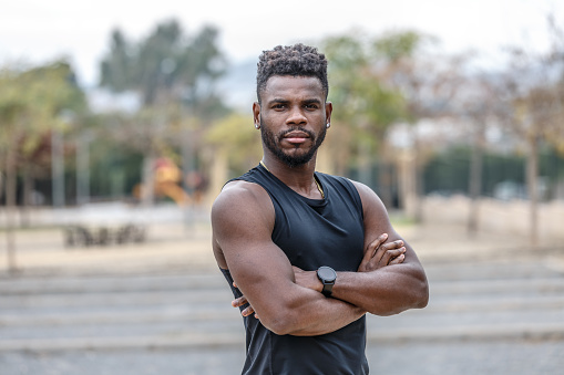 Muscular young black bearded male athlete with wristwatch standing on city street and crossing hands during fitness training