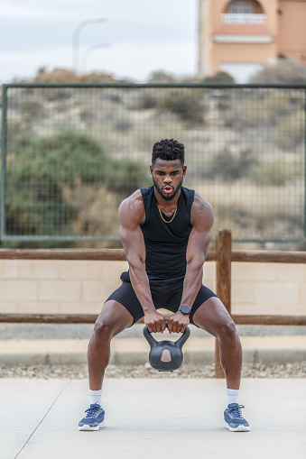 Full body of muscular African American male athlete lifting heavy kettlebell and taking deep breath during functional training