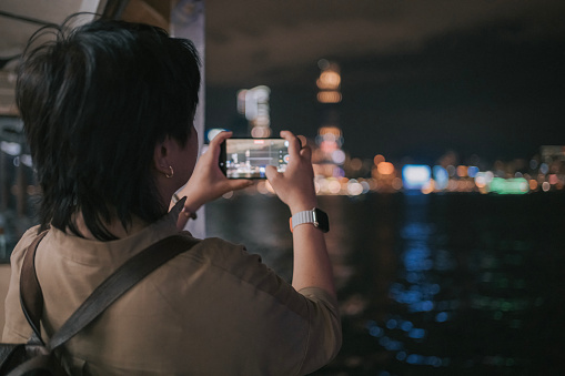 Asian Chinese female tourist filming hong kong cityscape at night taking star ferry crossing Hong Kong Island to Kowloon