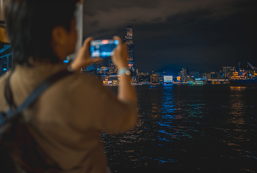 Asian Chinese female tourist filming hong kong cityscape at night taking star ferry crossing Hong Kong Island to Kowloon