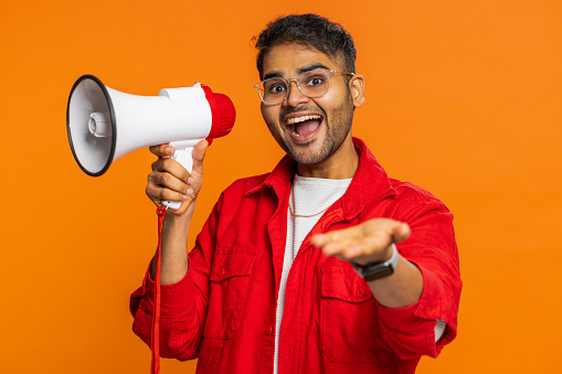 Indian Hindu man talking with megaphone, proclaiming news, loudly announcing advertisement discounts sale, using loudspeaker to shout speech. Happy Arabian guy isolated on studio orange background
