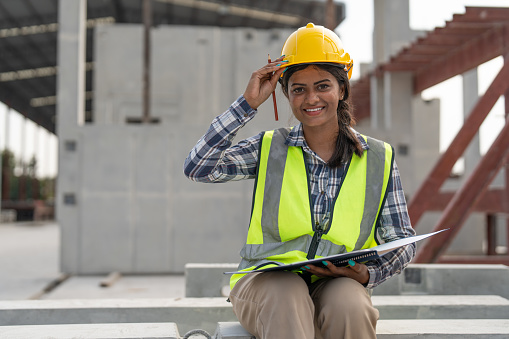 Asian woman engineer holding document smiling at construction site. Confident female Indian wearing protective helmet and vest setting in factory making precast concrete wall for real estate housing.