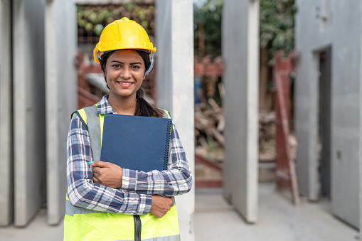 Asian woman engineer holding document smiling at construction site. Confident female Indian wearing protective helmet and vest working in factory making precast concrete wall for real estate housing.