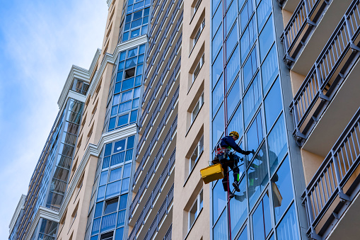 Bottom view of industrial mountaineer man laborer in work uniform washing glazing, hanging from building. Male rope access worker during high-rise job. Industry urban works concept. Copy ad text space