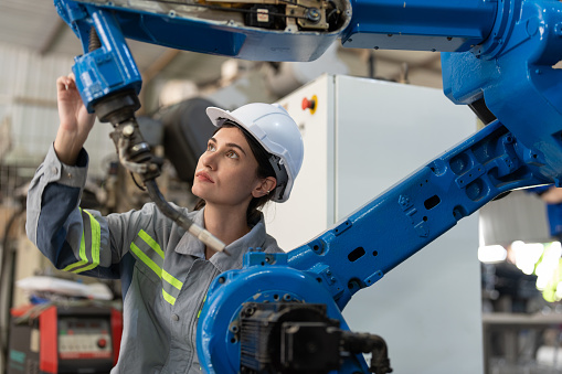 Female automation engineer wear vest with helmet safety checking and inspection control robot arm welding machine in an industrial factory. Artificial intelligence concept.