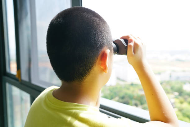 A young boy is playing with a toy car outdoors in front of a backdrop of tall buildings on a sunny day. He is wearing a brown and gray jacket and appears to be enjoying his playtime in the urban environment