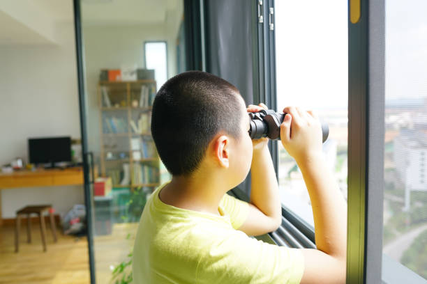 A young boy with short black hair is standing outdoors in a field holding a camera. He is wearing a brown long-sleeve vest and appears to be enjoying a casual moment in nature on a sunny day
