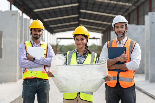 Group Indian construction team engineer wear vest and helmet safety and female leader hold blueprint standing together at factory construction site for making precast concrete wall.