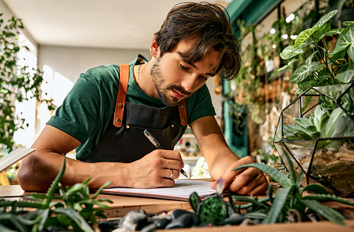 Focused caucasian man leaning on desk and writing on clipboard while working at modern store. Bearded florist running successful nursery with green domestic plant.