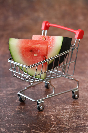 Stock photo showing close-up view of watermelon quarters sat in a miniature shopping trolley against a brown background. Healthy eating online shopping concept.