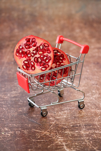 Stock photo showing close-up view of pomegranate (Punica granatum) halves sat in a miniature shopping trolley against a brown background. Healthy eating online shopping concept.
