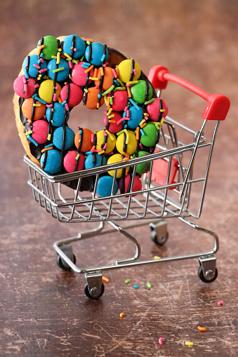 Stock photo showing close-up view of a glazed doughnut with a hole, decorated with chocolate drops covered with a hard candy coating in a miniature, model shopping trolley. Unhealthy eating sweet junk food concept.