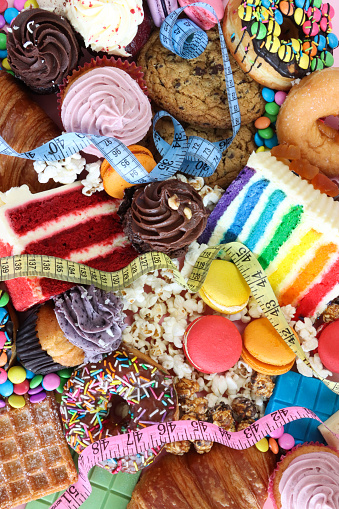 Stock photo showing close-up image of a variety of sweet junk food items including glazed ring doughnuts, popcorn, red velvet and rainbow cake slices, chocolate chip cookies, cupcakes with butter icing, chocolate bars, macarons, waffle, croissants. Unhealthy eating concept.