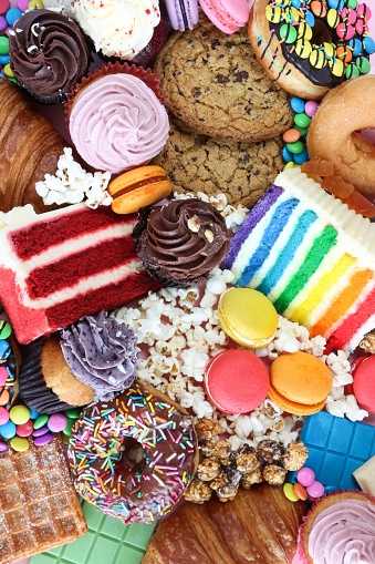 Stock photo showing close-up image of a variety of sweet junk food items including glazed ring doughnuts, popcorn, red velvet and rainbow cake slices, chocolate chip cookies, cupcakes with butter icing, chocolate bars, macarons, waffle, croissants, against a pink background. Unhealthy eating concept.