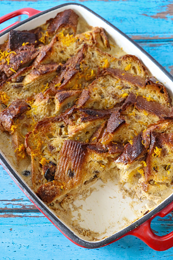 Stock photo showing elevated view of the layered ingredients for a bread and butter pudding with a missing portion in a red baking dish on blue wood grain background.