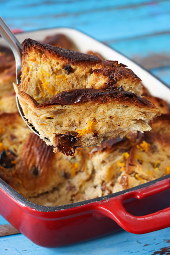 Stock photo showing close-up, elevated view of the layered ingredients for a bread and butter pudding in a red baking dish on blue wood grain background.