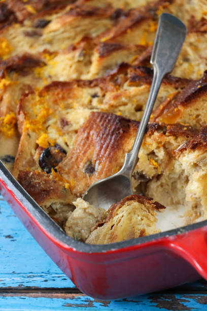 Stock photo showing close-up, elevated view of the layered ingredients for a bread and butter pudding in a red baking dish on blue wood grain background. close-up image of red baking dish containing cooked ingredients of layered bread and butter pudding, slices of buttered white bread layers sprinkled with dried fruit, metal spoon, blue wood grain background, elevated view, focus on foreground