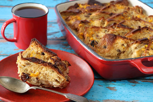 Stock photo showing close-up view of a bread and butter pudding in a red baking dish besides serving of dessert on red plate with metal spoon and a red mug of black coffee.
