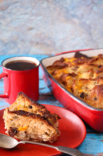 Stock photo showing close-up view of a bread and butter pudding in a red baking dish besides serving of dessert on red plate with metal spoon and a red mug of black coffee.