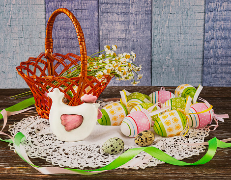 Easter composition with a basket with Easter eggs, decorative chicken, quail eggs on a wooden background. Chamomile flowers, feathers and green ribbon as decor.