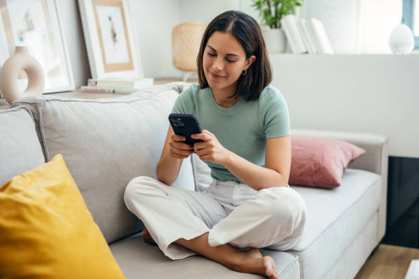 istock Smiling young woman using her mobile phone while sitting on sofa at home 1911998815