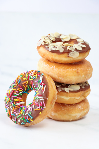 Stock photo showing close-up view of single chocolate glazed ring doughnut, topped with white, pink, green, blue, yellow, and orange, hundred and thousand sugar sprinkles leaning against tower of plain and chocolate glazed deep fried desserts.