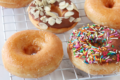 Stock photo showing close-up view of rows of alternating plain and chocolate glazed ring doughnuts on cooling rack. Some of the chocolate glazed cakes are topped with flaked almonds, whilst others are decorated with white, pink, green, blue, yellow, and orange, hundred and thousand sugar sprinkles.